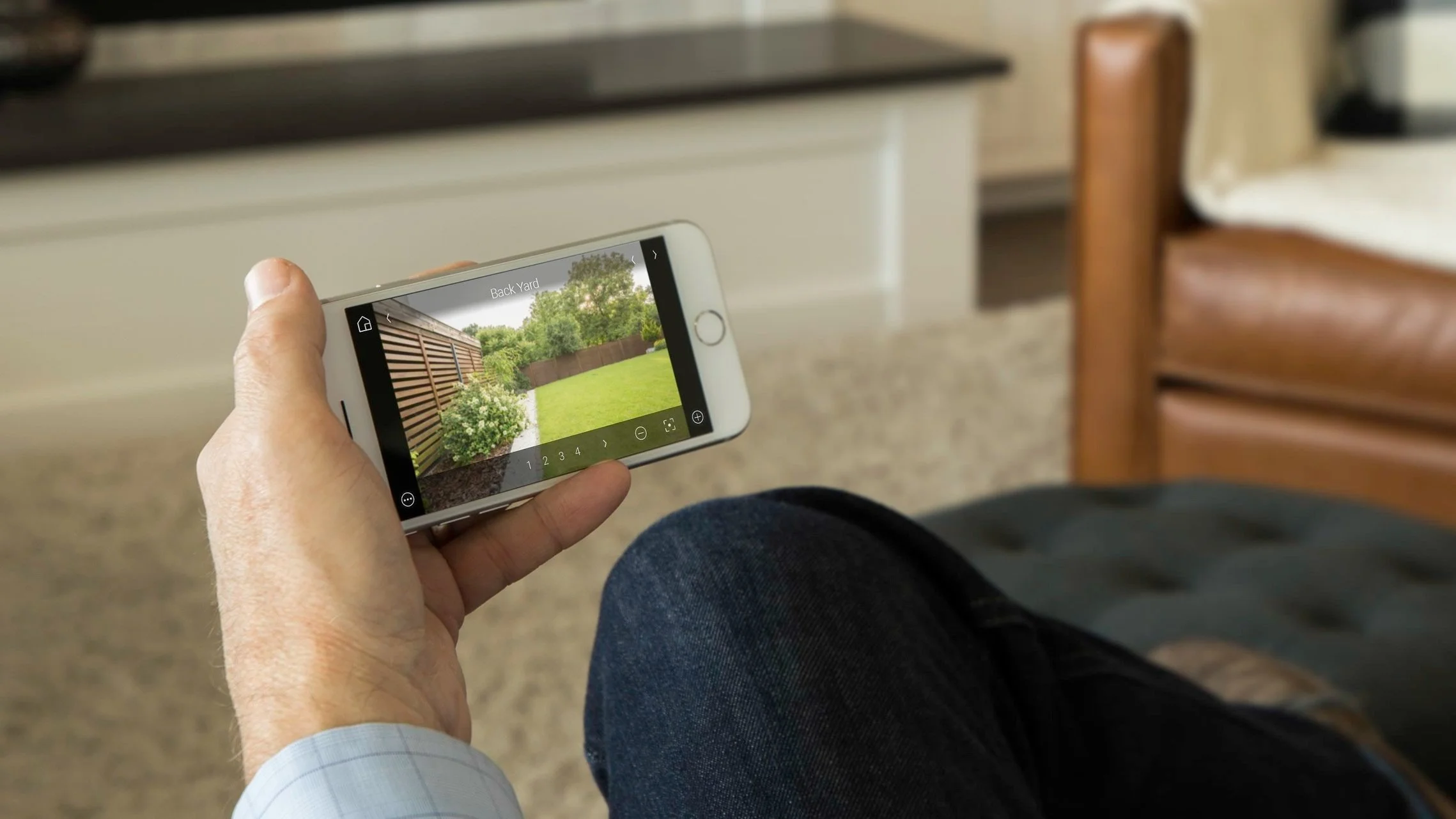 Person sitting on a chair holding a smartphone, viewing a photo of a backyard with a lawn, trees, and a wooden fence.