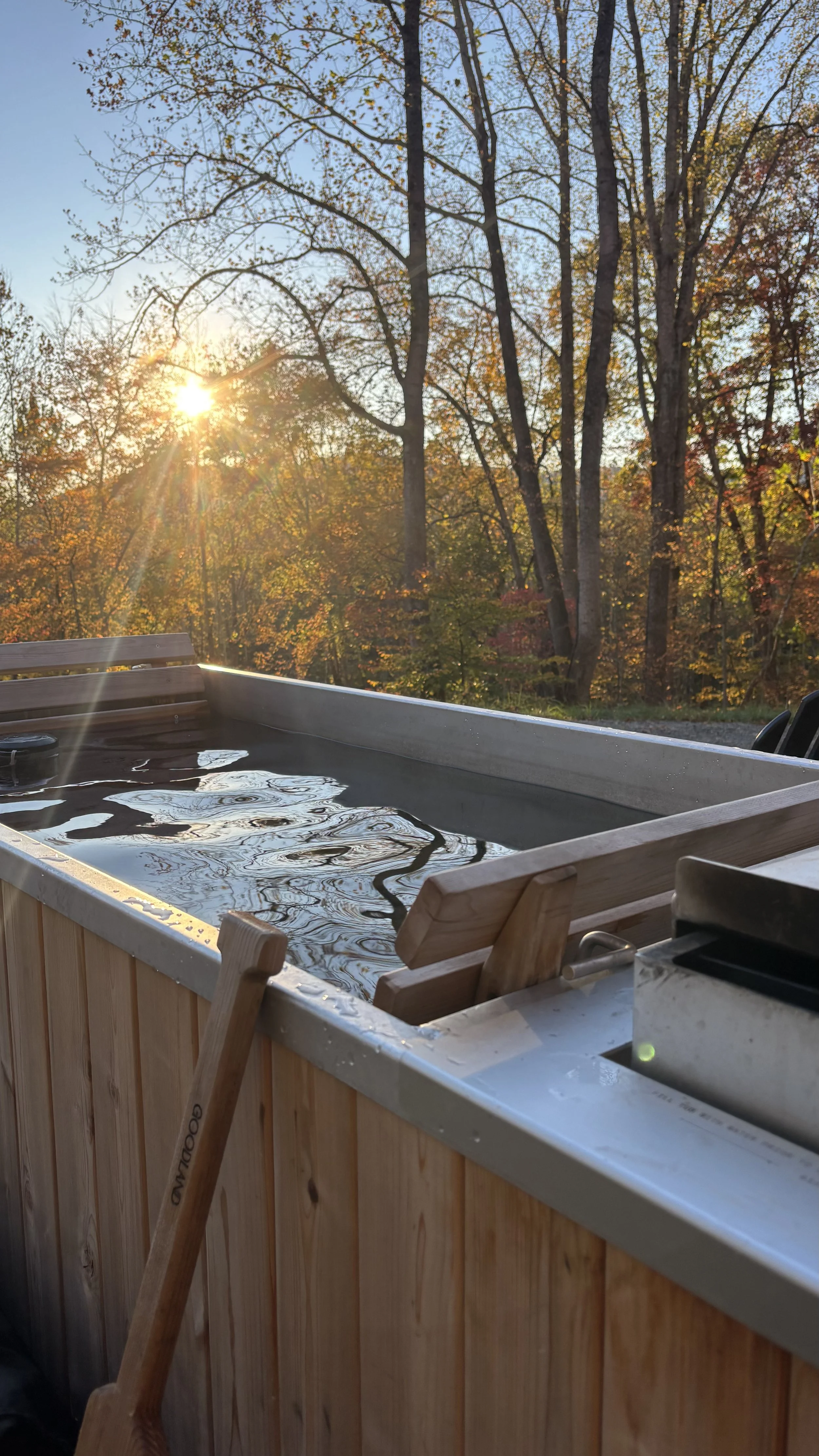 An outdoor hot tub with wooden sides, surrounded by chairs, set against a backdrop of trees with autumn foliage, during sunset.