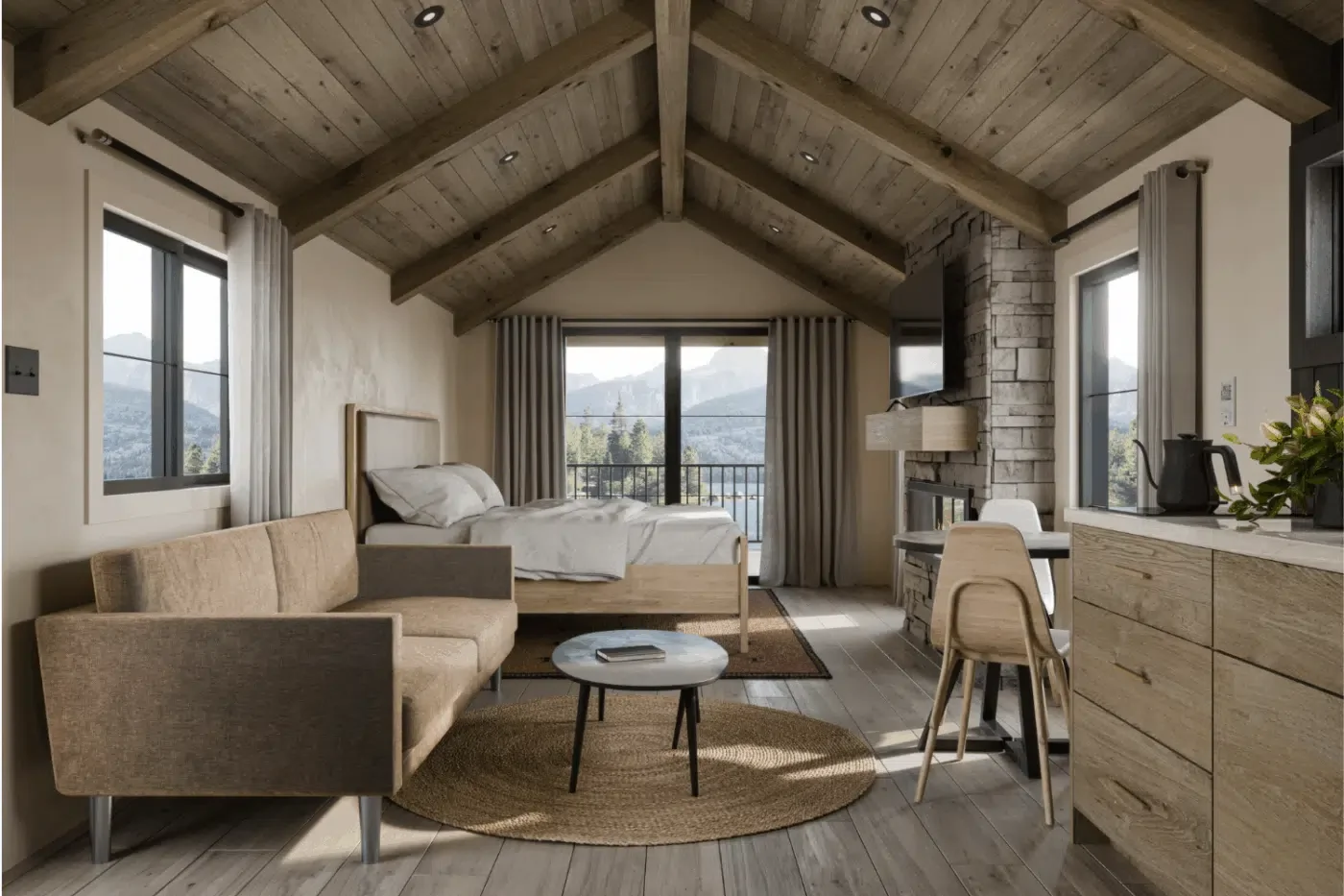 Interior of a cozy mountain lodge room with a vaulted wooden ceiling, large windows, a bed, a beige sofa, a small round coffee table, a stone fireplace, and wooden furniture, with mountains visible outside.
