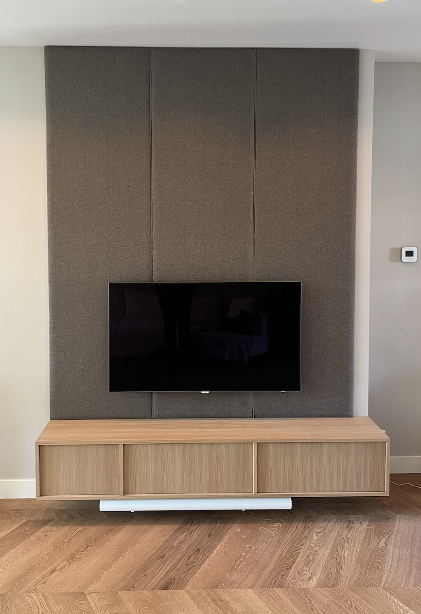 Een houten tv-meubel met een bekleedde wandpaneel achter de tv. Op maat gemaakte textiele wandmeubel. Maatwerk Den Haag. 