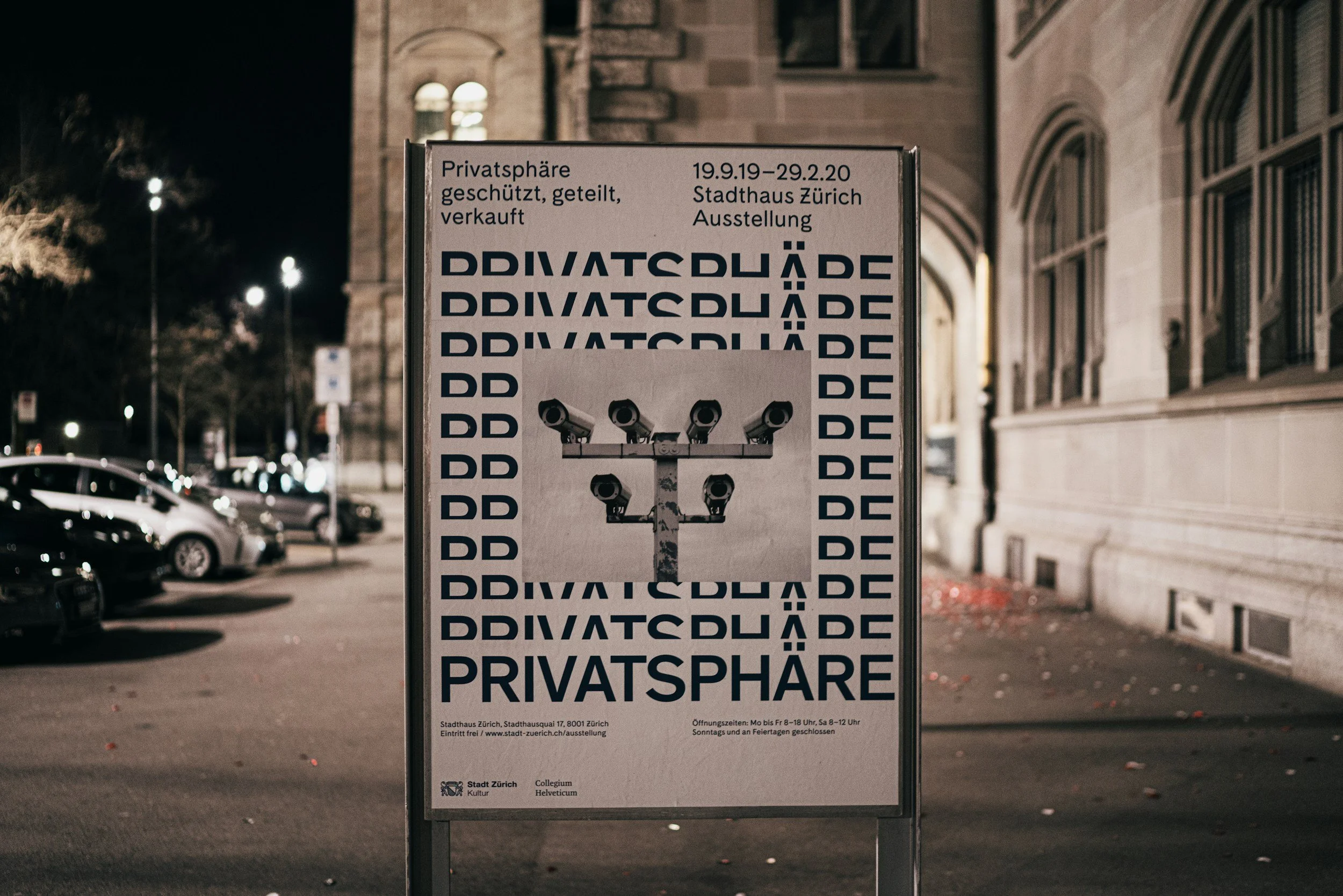 A sign standing outside a building at night, advertising an exhibition at the Zurich City Hall from September 19, 2019, to February 29, 2020, with an illustration of multiple security cameras.