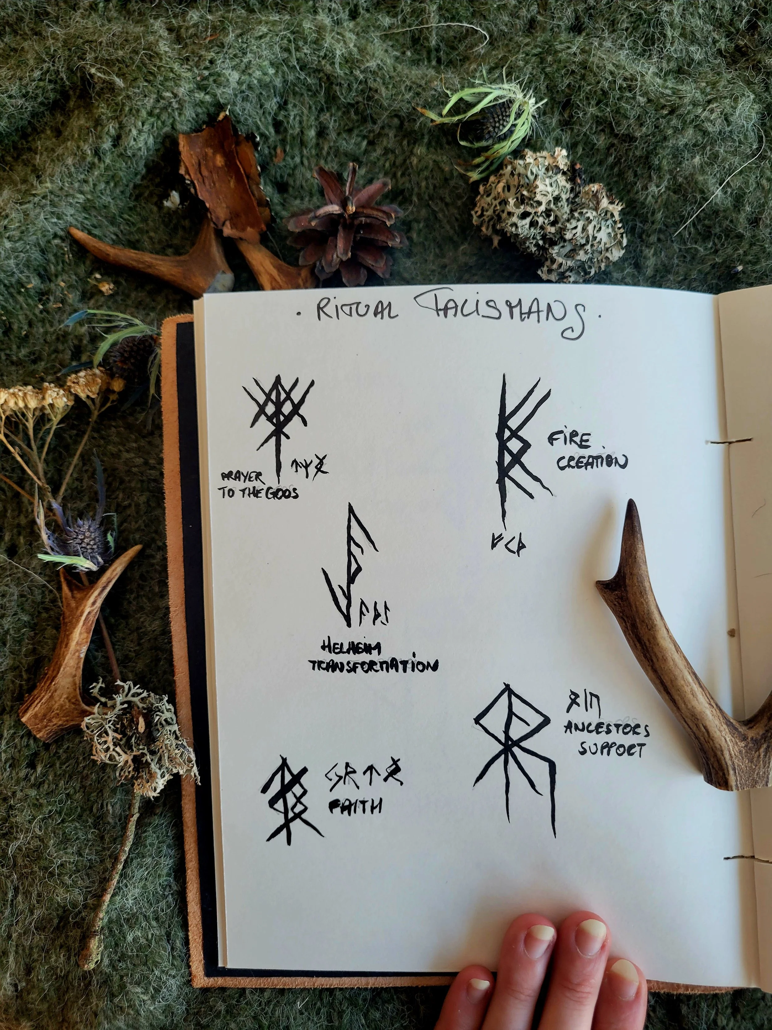 A notebook page with various symbols and words related to ritual practices, including phrases like 'prayer to the gods,' 'fire creation,' 'hellism transformation,' and 'ancestral support.' Surrounding the notebook are natural objects such as pinecones, bark, dried flowers, and lichen, all placed on a textured green surface.