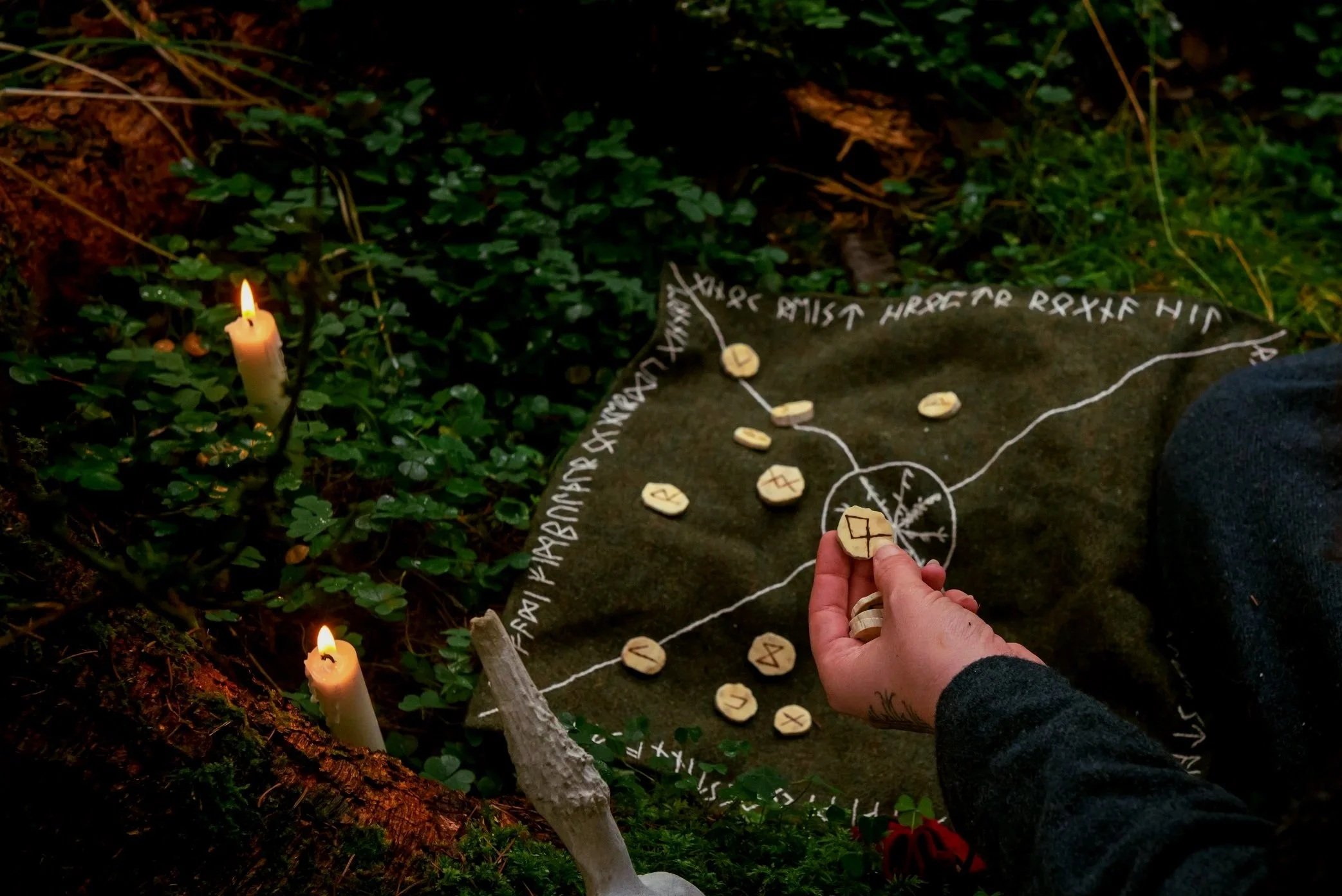 Person holding runes over an outdoor rune chart with candles and green plants surrounding them.