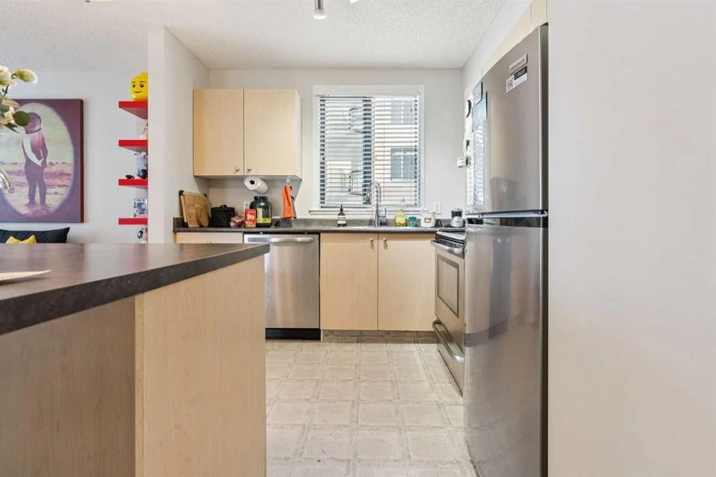 A kitchen with light wooden cabinets, a stainless steel refrigerator, and a window above the sink with blinds.