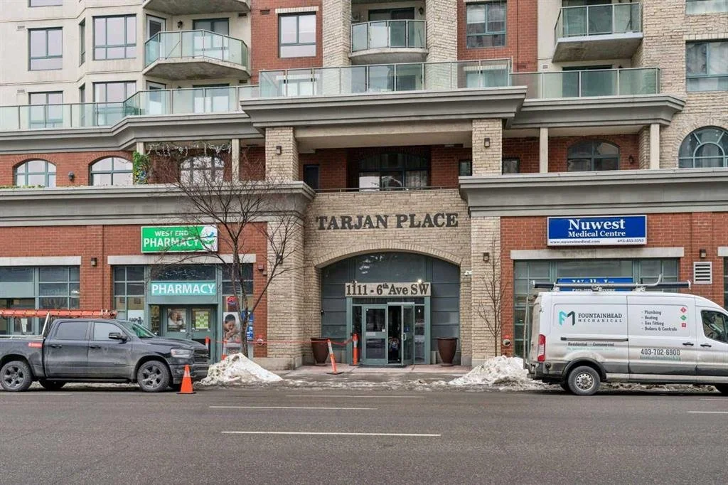 Street view of a multi-story residential building with a bank named Tarjan Place, a pharmacy, and a medical center entrance. cars parked in front and some snow on the sidewalk.