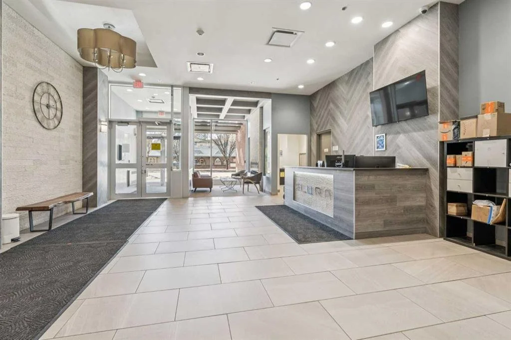 Lobby with reception desk, large wall clock, and seating area with chairs and coffee table. Entrance with glass doors, and a hallway with a window showing trees outside.