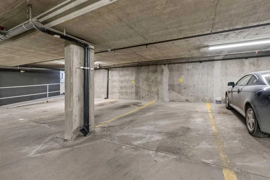 Empty parking garage with one car parked in a space on the right. Concrete walls, ceiling, and floor with yellow parking space lines and institutional lighting.
