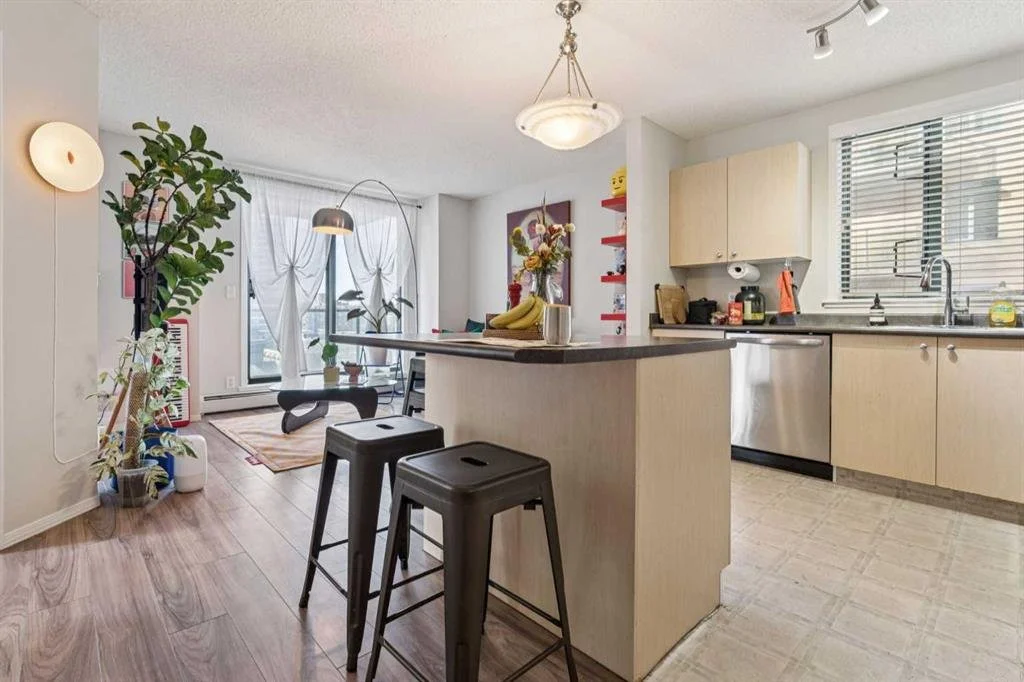 Open kitchen and living room with a small kitchen island, two black bar stools, a large window with blinds, potted plants, wall art, and a round ceiling light.
