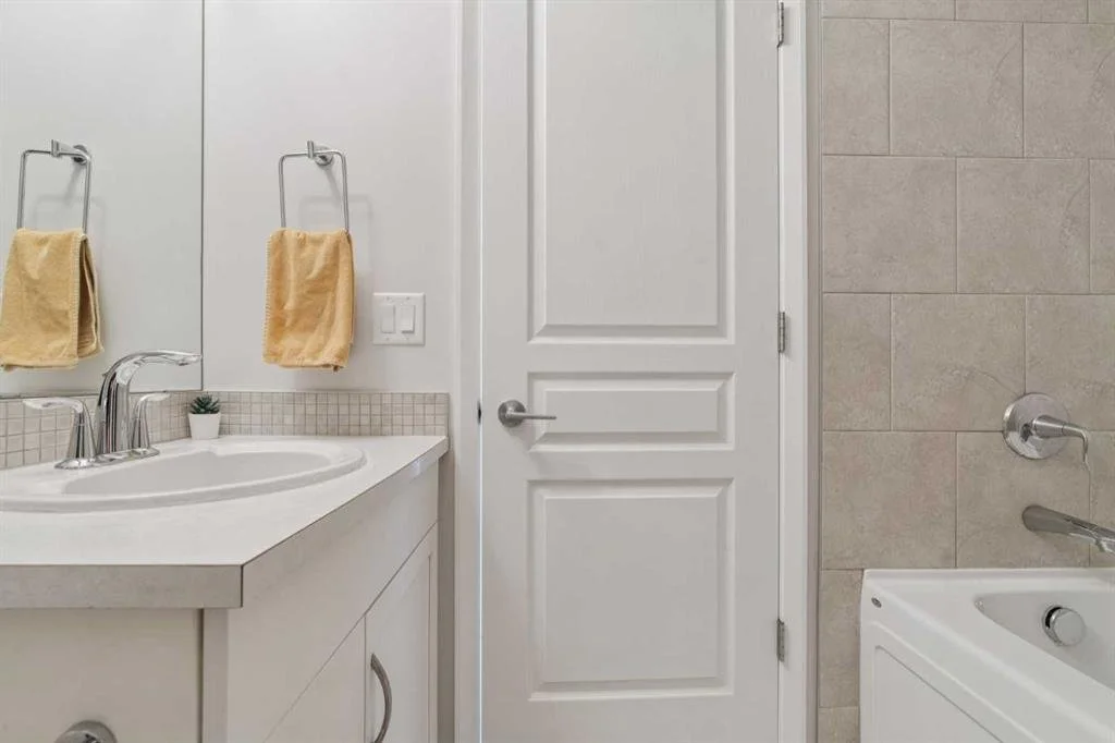 Bathroom with a white vanity, a small potted plant, beige towels on towel racks, a door, and part of a bathtub with a faucet and tile wall.