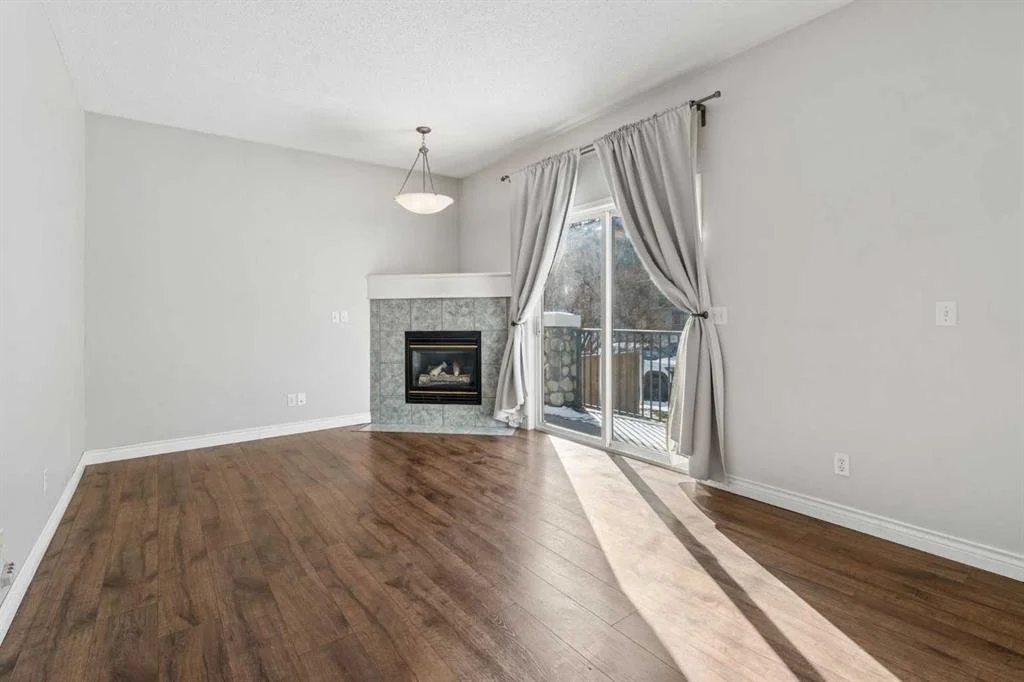 Empty living room with hardwood floors, white walls, a sliding glass door with curtains leading to a balcony, and a fireplace with tiled surround.