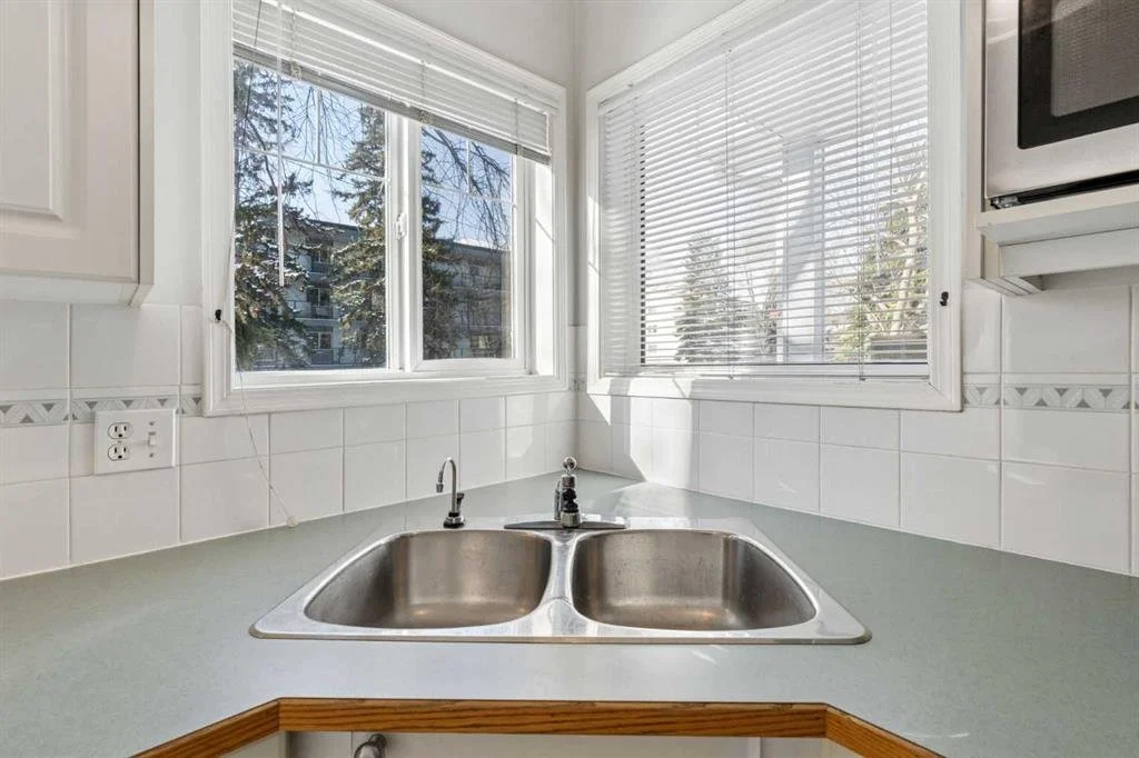 Kitchen sink area with two basins, white tiled backsplash, and three windows with blinds overlooking trees.