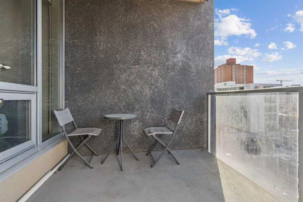 Small balcony with a round table and two folding chairs, with a large textured wall and glass railing, overlooking city buildings and a partly cloudy sky.