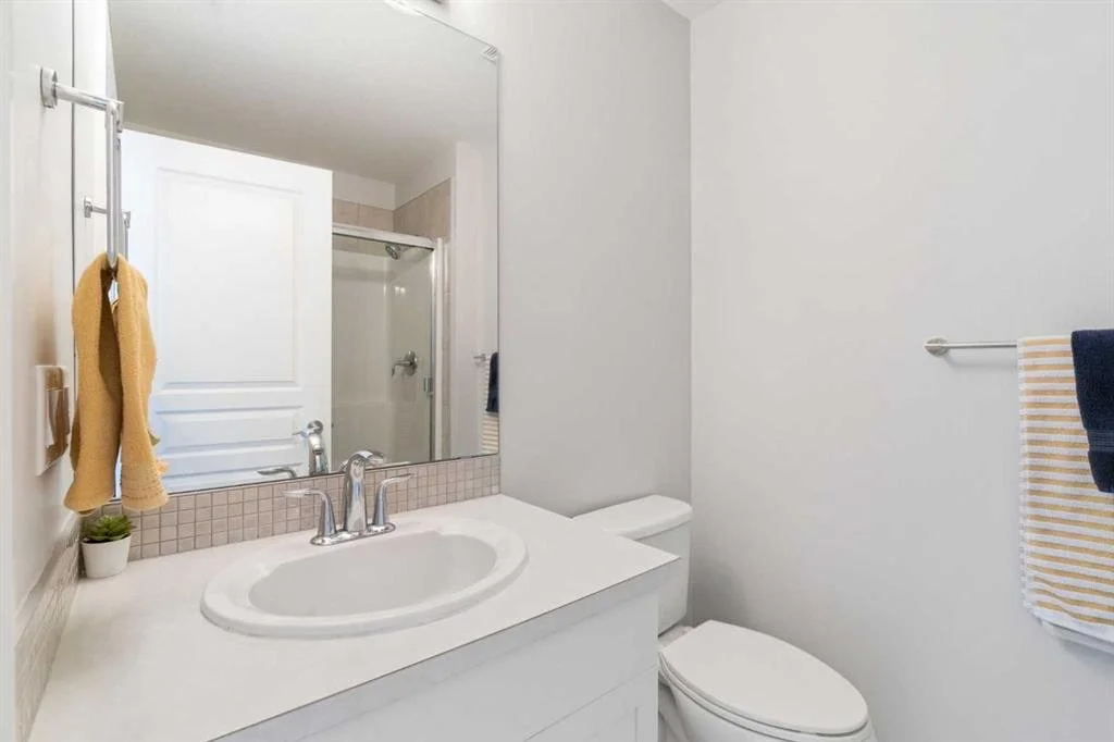 A small modern bathroom with a white sink, a mirror above, a small plant on the counter, a towel rack with yellow and striped towels, and a white toilet. The shower has a glass door and is visible behind the mirror.