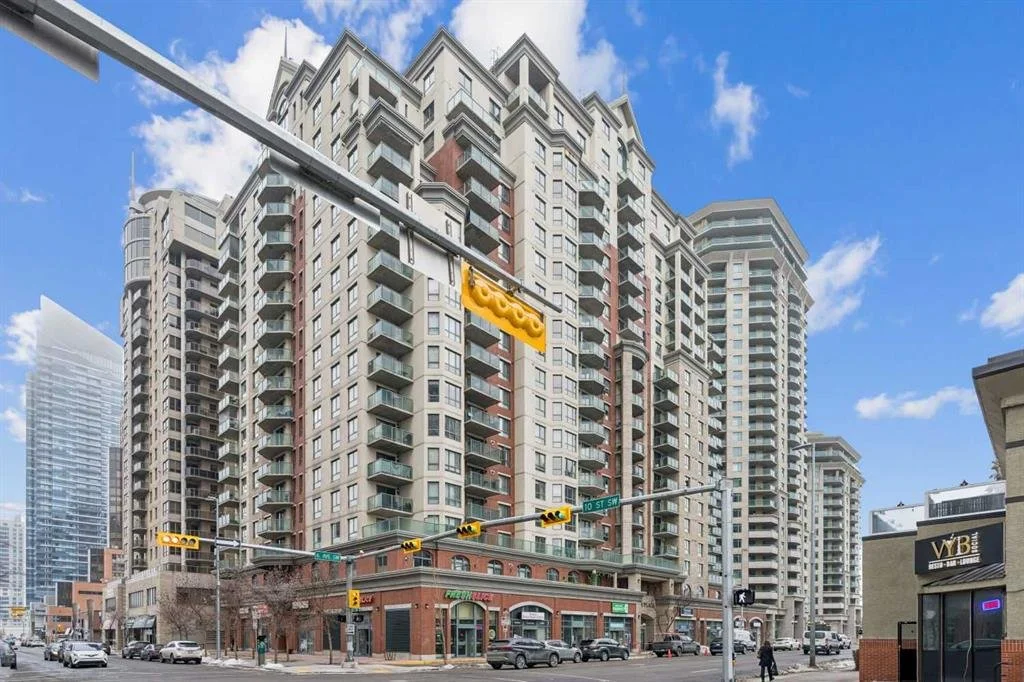 A tall multi-story apartment building with multiple balconies, situated at a street corner with traffic lights and several cars parked on the street. The building has a mix of modern and classic architectural elements, with a commercial space on the 