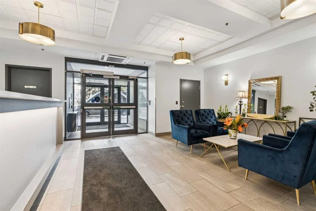 Hotel lobby with gray doors and glass entrance, cream-colored walls, modern gold and white light fixtures, a seating area with two navy blue velvet armchairs and a sofa, a white coffee table with flowers, a gold-framed mirror, and a console table wit