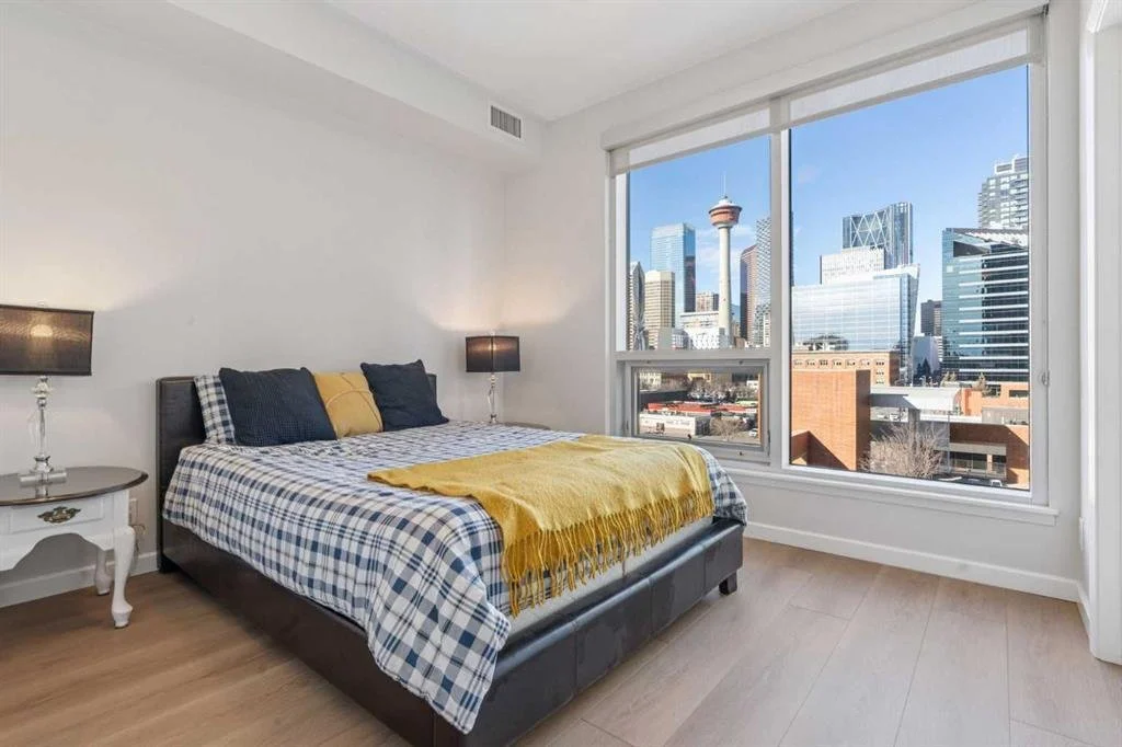 Bedroom with a large window showing a city skyline with tall buildings and a tower, bed with plaid bedding, yellow blanket, and dark pillows, beside a white nightstand with a lamp.