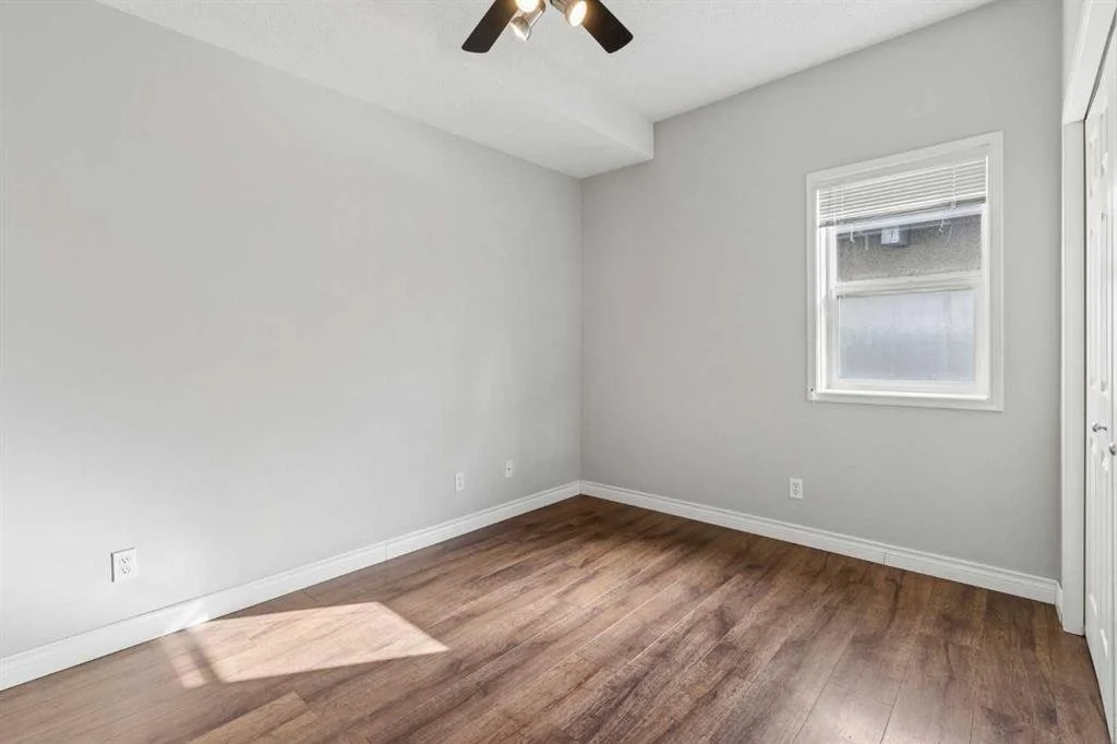 Empty bedroom with white walls, a window with blinds, a closet with sliding doors, wood flooring, and a ceiling fan with lights.