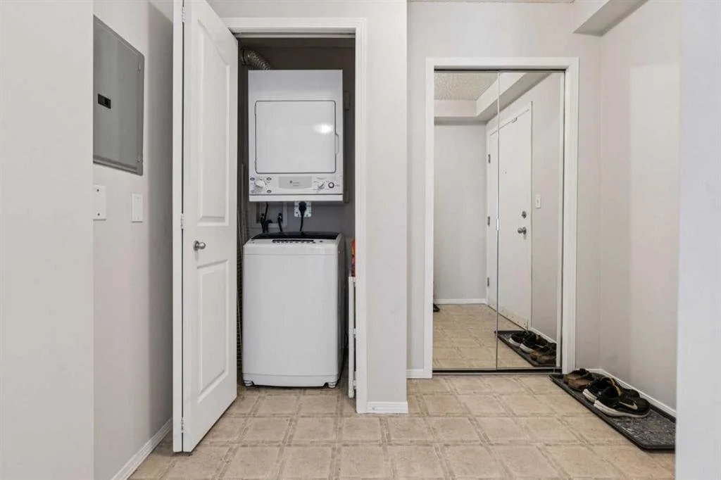 A laundry area with a stacked washer and dryer in a small closet, a mirror wardrobe, and multiple pairs of shoes on mats near the entrance.