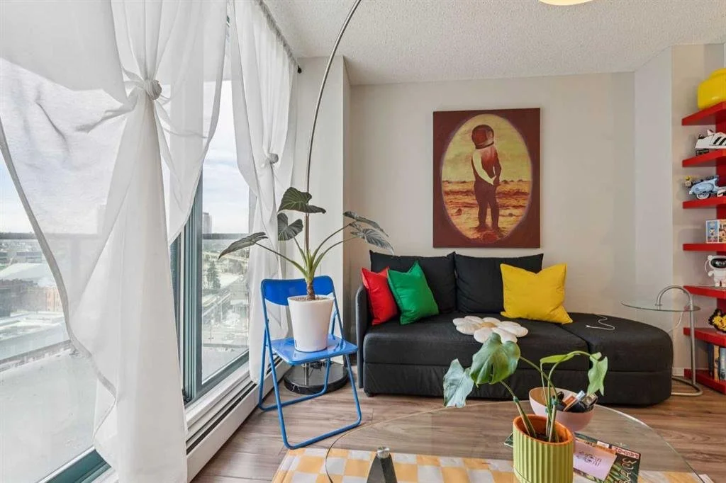 Living room with a large window with sheer white curtains, a black sofa with multicolored pillows, a painting of a child with a dog on the wall, a blue chair with a potted plant, and a red shelf with toys.