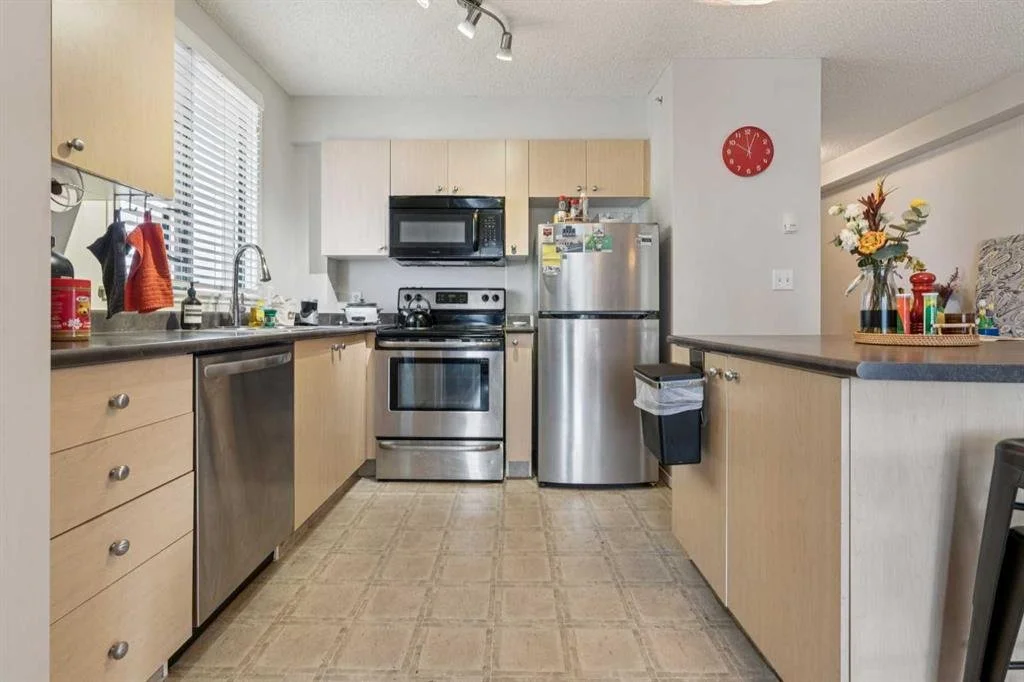 A kitchen with light wood cabinets, stainless steel appliances, including a fridge, stove, microwave, and dishwasher. There is a window with blinds above the sink, and various items on the counters, including a flower vase, containers, and cleaning s