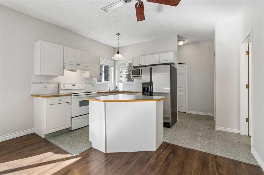 White kitchen with cabinetry, microwave, stove, refrigerator, and small island, with a wood and tile floor, and a ceiling fan.