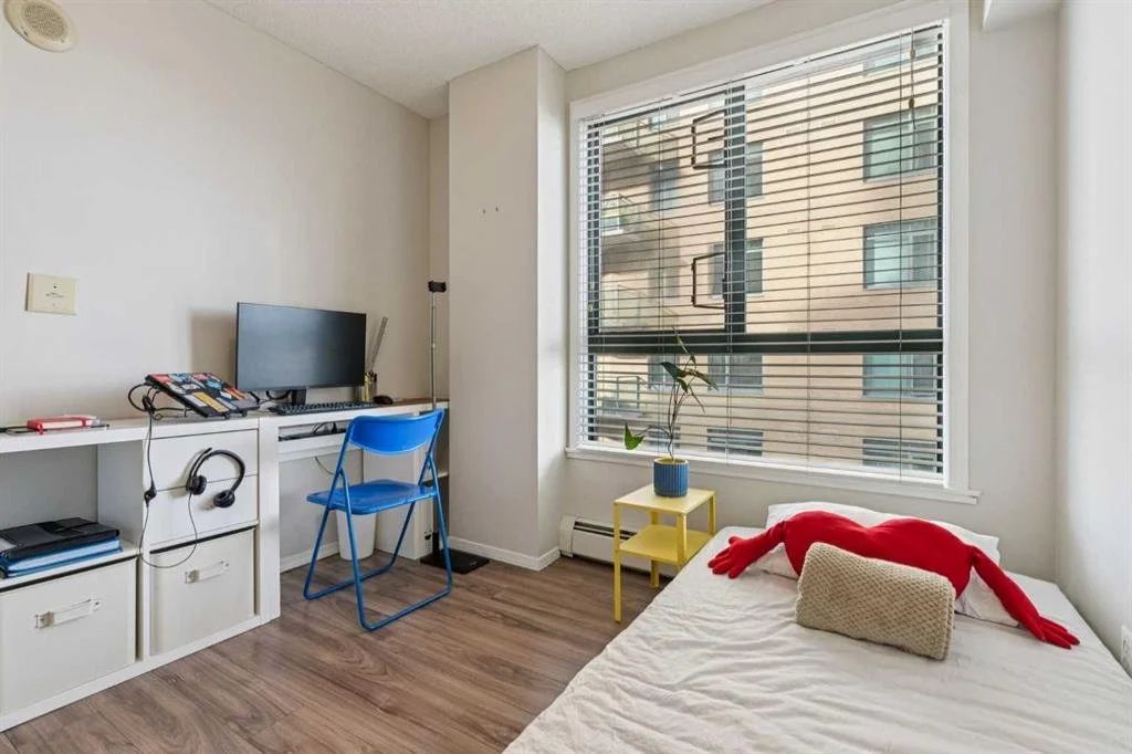 A bedroom with a window, a bed with a plush red toy, a small yellow side table with a potted plant, and a white desk with a computer, keyboard, and headphones. The room has wooden flooring and white walls.