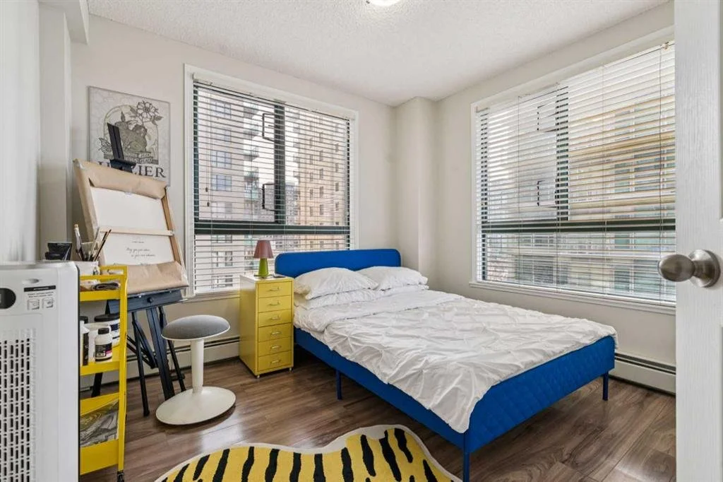 A bedroom with a blue bed, yellow nightstand, and large windows with blinds. A white chair, a small lamp, and a yellow shelving unit are near the bed, and hardwood floors are visible.