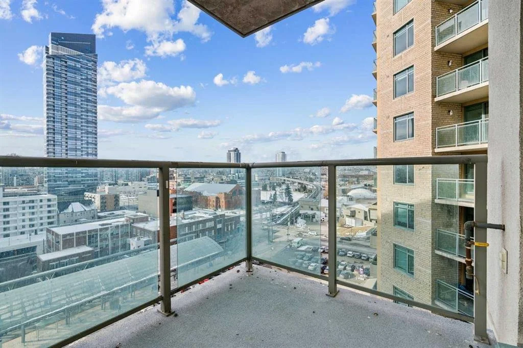City skyline view from an apartment balcony with glass railing, neighboring buildings, and elevated train tracks under a partly cloudy sky.