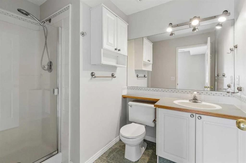 Small bathroom with a shower on the left, a toilet, and a sink with a large mirror and lighting fixtures above. White cabinets and a partial wall with decorative tiles.