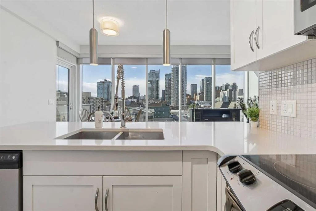 Modern kitchen with white cabinetry, a sink with a spring faucet, and a view of city buildings through large windows, with pendant lights hanging from the ceiling.