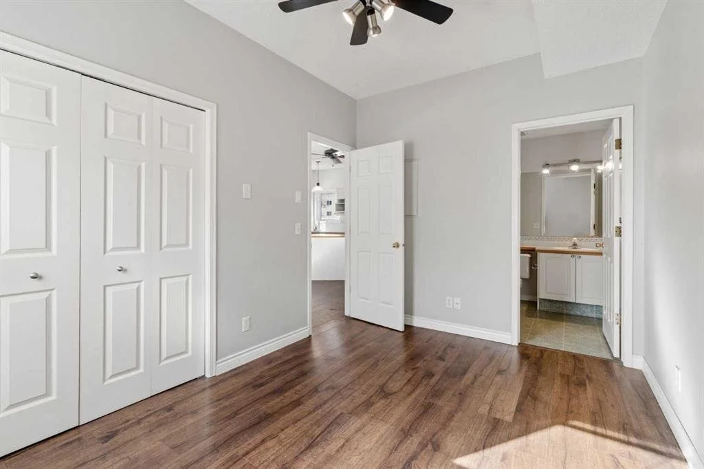 Empty bedroom with light gray walls, white trim, hardwood floor, closet with white bifold doors, and an open door leading to a bathroom with white cabinets and a mirror.