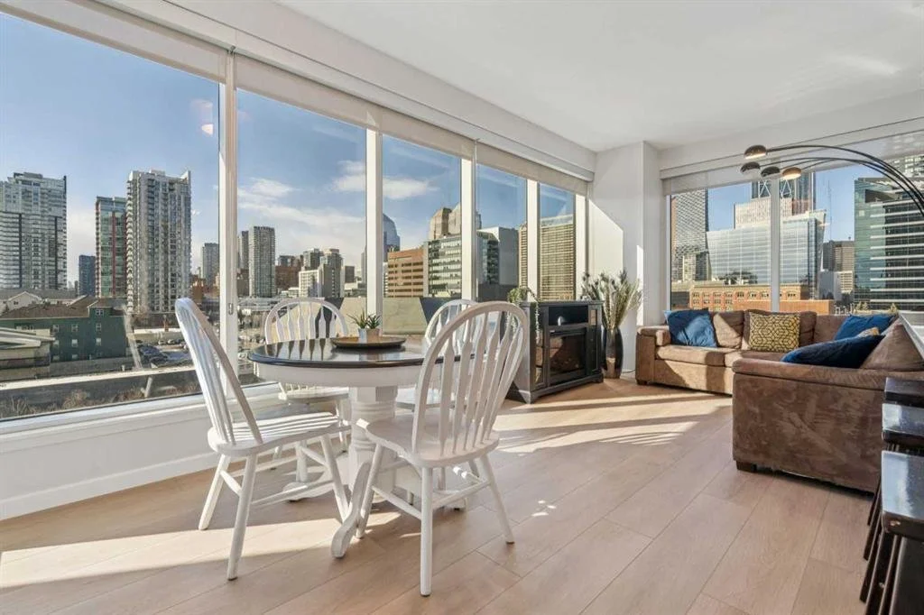 Sunlit apartment living room and dining area with floor-to-ceiling windows showing a city skyline, brown sofa with colorful pillows, round dining table with white chairs, and a black floor lamp.