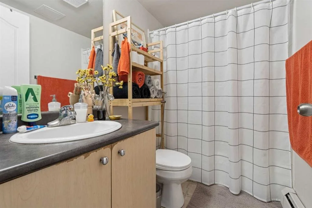 A bathroom with a countertop sink, a mirror, and a shower with a white curtain. The counter has toiletries and decor items, and there are colorful towels and a foldable wooden shelving unit with towels and clothes.