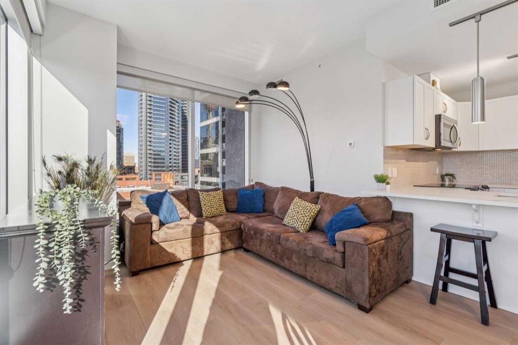 Living room with a large brown sectional sofa, blue and yellow pillows, a floor lamp with multiple adjustable heads, and large windows showing city buildings, with a kitchen counter and appliances in the background.