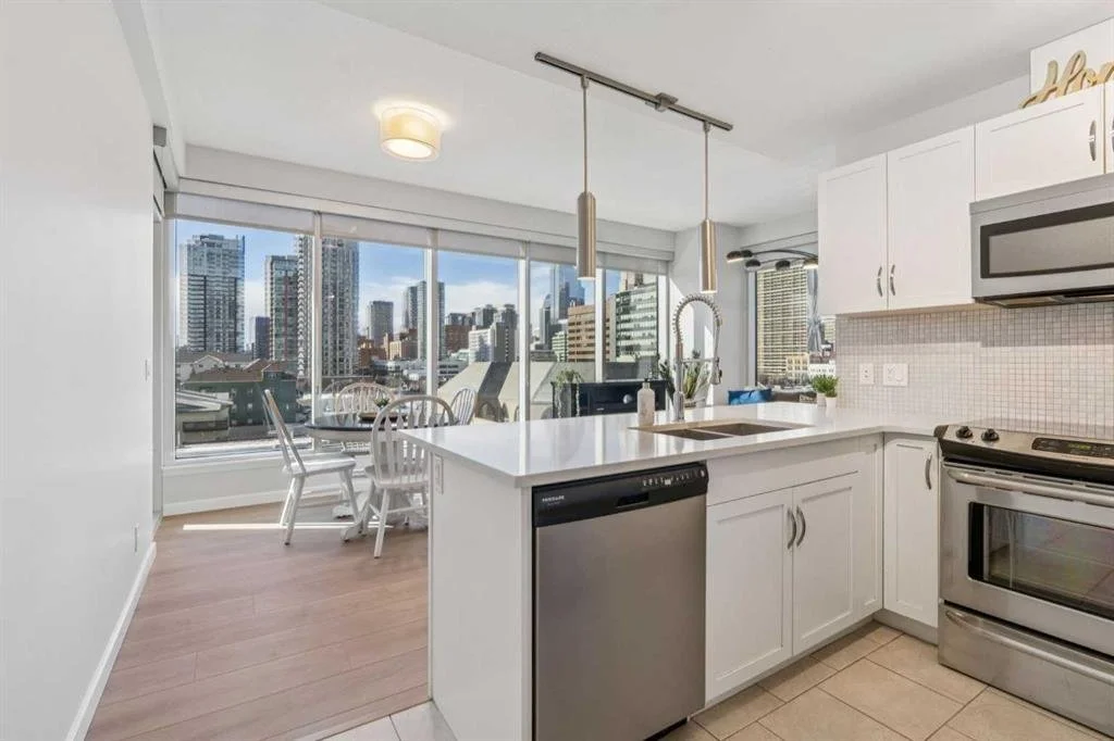 Modern kitchen with white cabinetry and stainless steel appliances, overlooking city skyline through large windows, with a dining area in the corner.