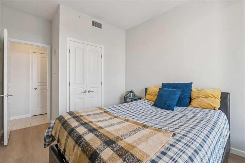 A bedroom with a bed covered in a plaid and checkered blanket, blue and yellow pillows, a small side table with plants next to the bed, and a closed white closet door. Adjacent to the room, an open door reveals a hallway with another door.