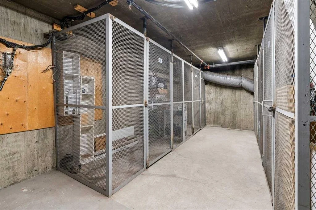 A utility or storage room with cement walls and floor, enclosed metal cages, electrical panels, and a large duct running along the ceiling.