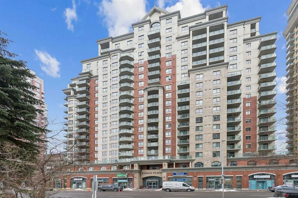 Tall multi-story residential building with balconies, retail shops on the ground level, and a clear blue sky above.
