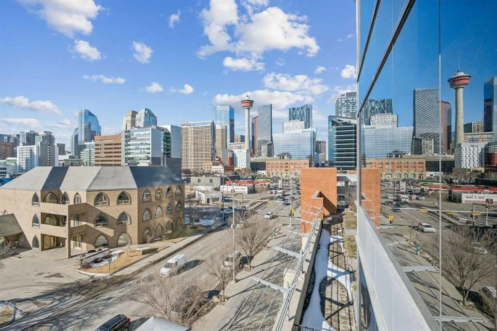 City skyline with various tall modern buildings, a tower with a circular observation deck, and a clear blue sky with scattered clouds, reflected on the glass windows of a building.