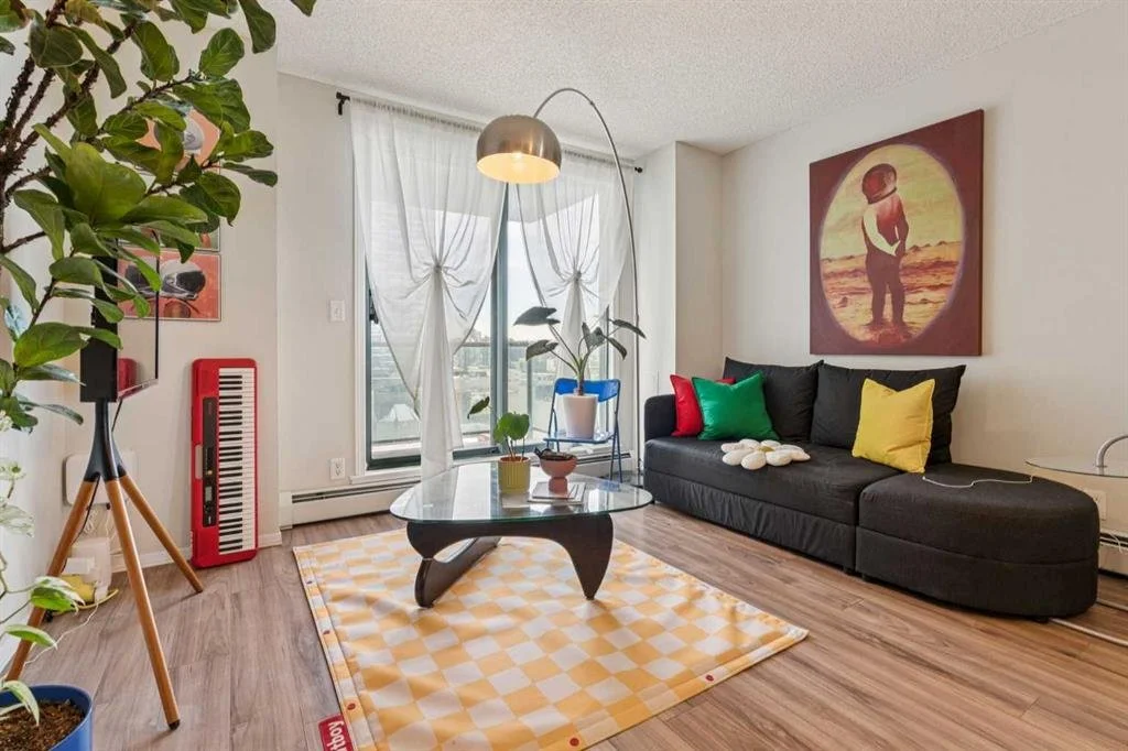 A modern living room with a black sofa with colorful pillows, a glass coffee table on a yellow checkered rug, a potted plant, a red keyboard, a large painting of a person at the beach, and a floor lamp near a sliding door with sheer curtains.