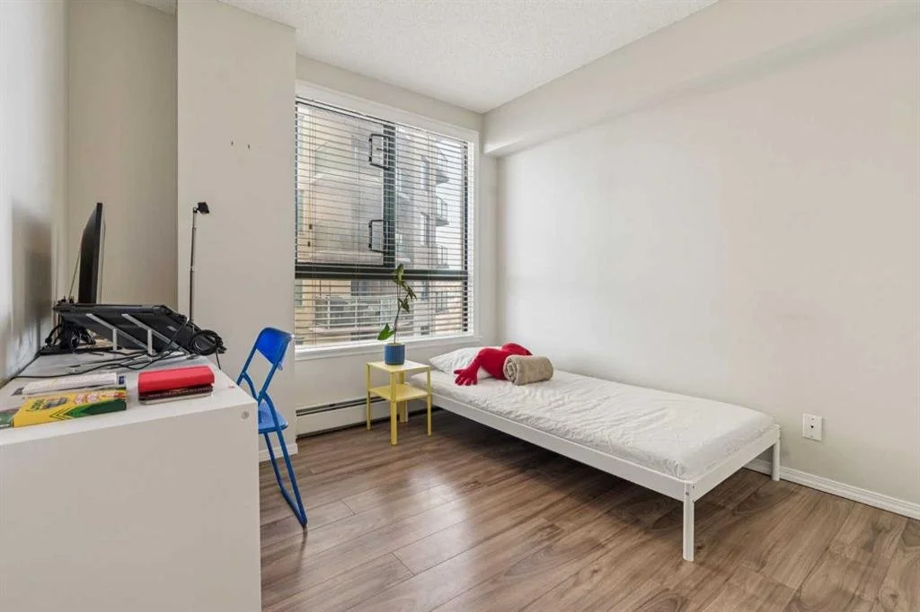 A minimalist bedroom with a single bed, a small yellow nightstand with a potted plant, a window with blinds, a blue chair, and a white desk with a computer monitor and some books and office supplies.