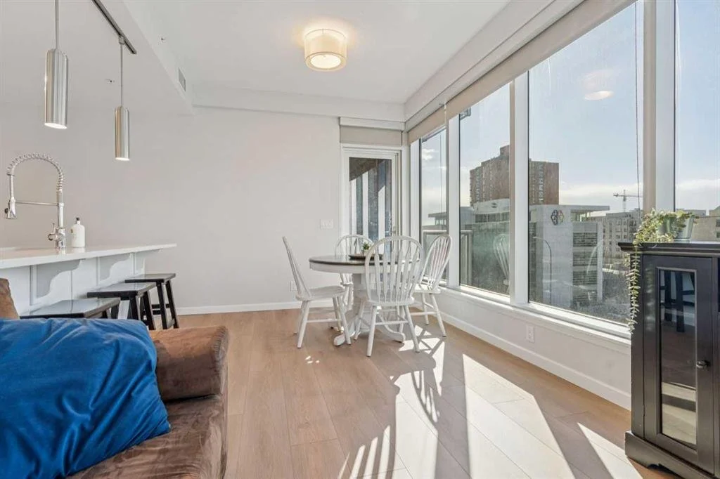 Sunlit apartment dining area with a round table and white chairs, large windows with city views, and a corner cabinet.