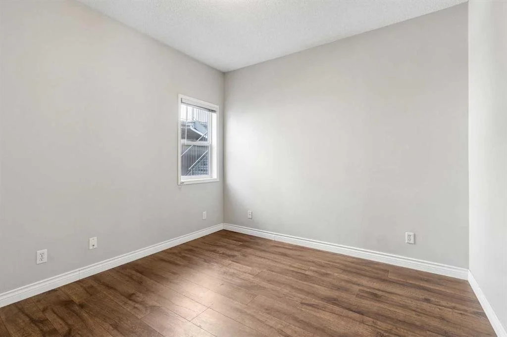 Empty room with white painted walls, a window showing an outdoor staircase, wooden flooring, and white baseboards.