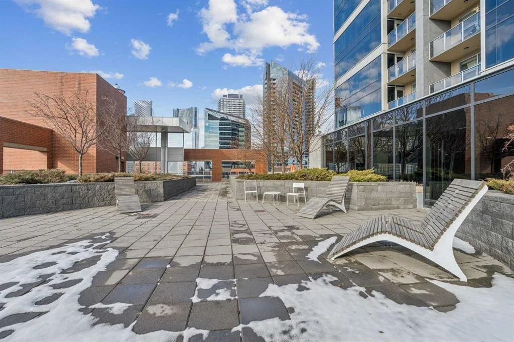 Empty outdoor terrace with modern lounge chairs, small tables, snow patches, and surrounding buildings under a partly cloudy sky.