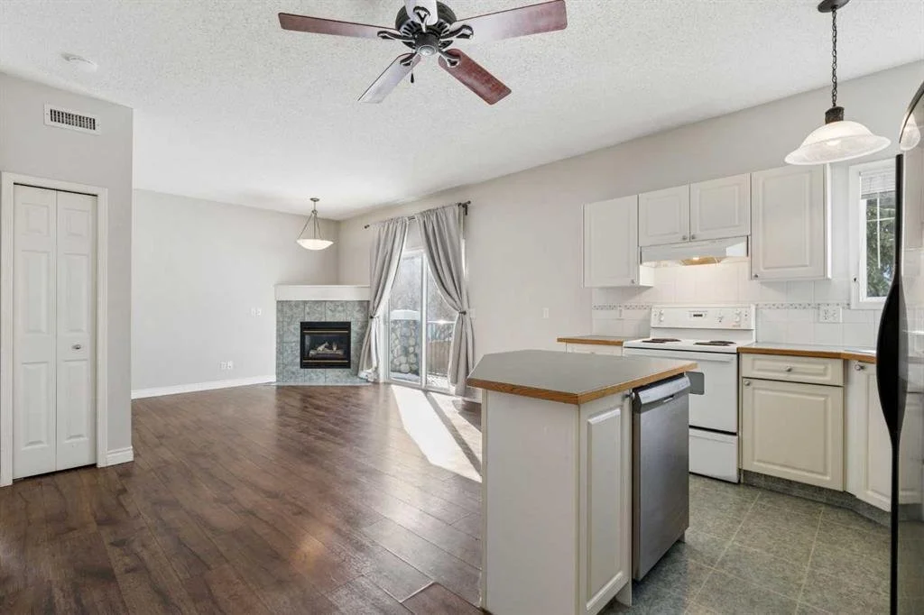 Open-concept living room and kitchen with white cabinetry, a fireplace, hardwood flooring, a ceiling fan, and a sliding glass door with curtains leading outside.