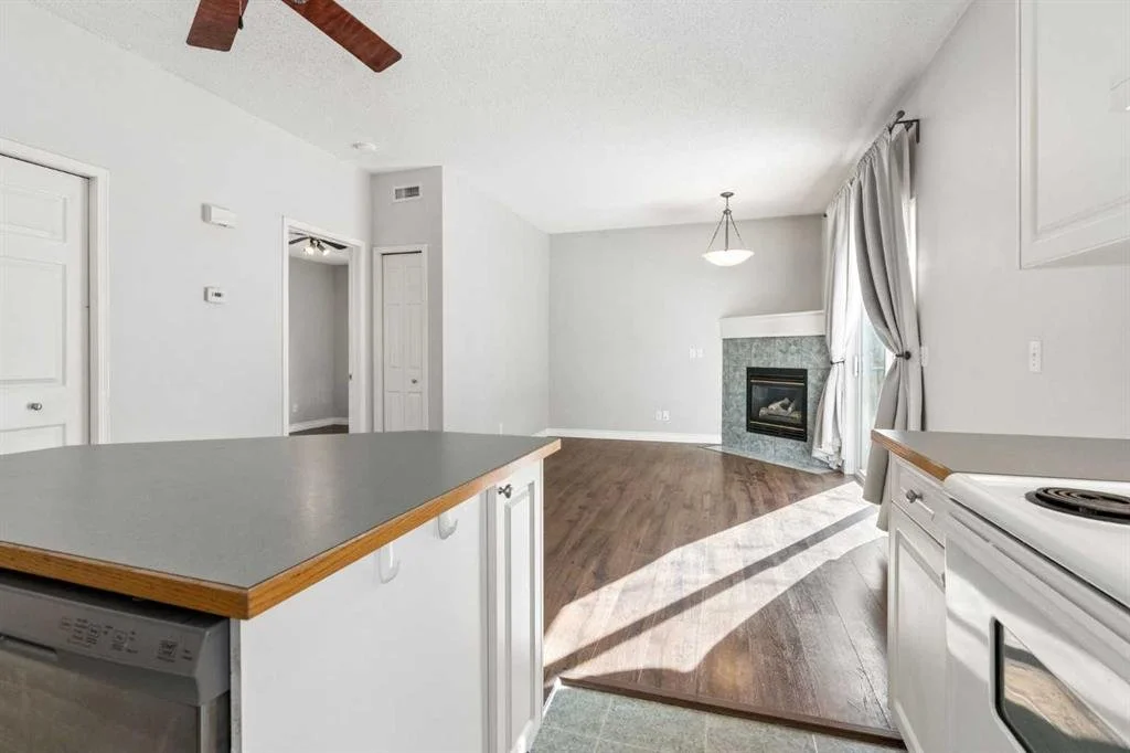 Empty living room with a corner fireplace, hardwood floors, large window with curtains, and an open kitchen with a countertop.