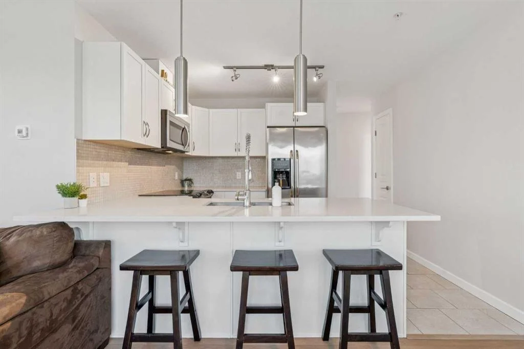 Modern kitchen with white cabinets, stainless steel refrigerator and microwave, granite countertops, small backsplash, and three dark stools at a breakfast bar.