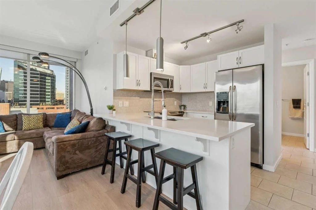 Open-concept kitchen and living room with large window showing cityscape, white cabinets, stainless steel refrigerator, and a brown sectional sofa with colorful pillows.