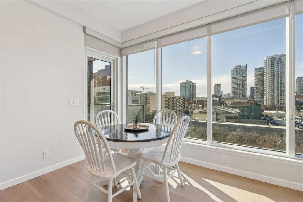 A round dining table with four white chairs in a bright corner with large floor-to-ceiling windows overlooking a cityscape with tall buildings.