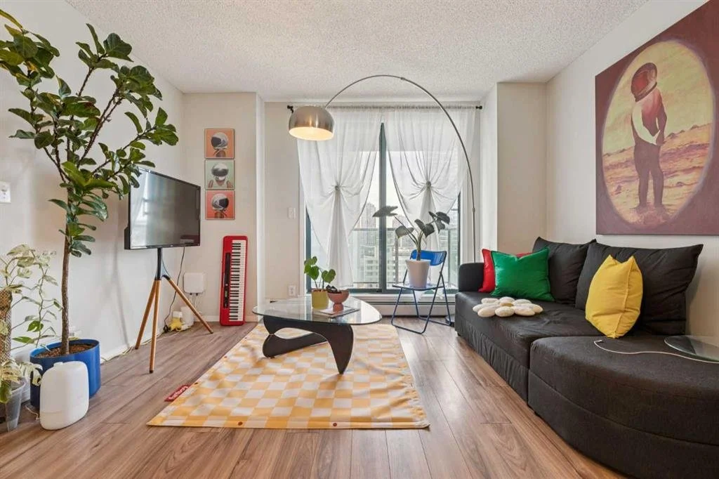 Living room with a large black sofa with colorful pillows, a yellow and white checkered rug, a modern coffee table, potted plants, a wall-mounted TV, wall art, and a window with sheer white curtains.